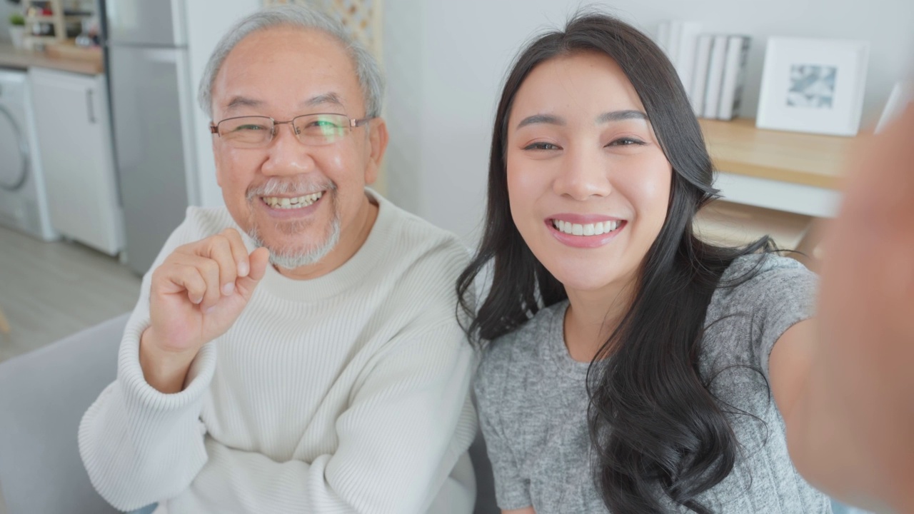 亚洲可爱的家庭，女儿用电话与资深父亲视频通话。年轻漂亮的女孩和上了年纪的老男人坐在沙发上享受网上交流，在家里的客厅里一起聊天视频素材