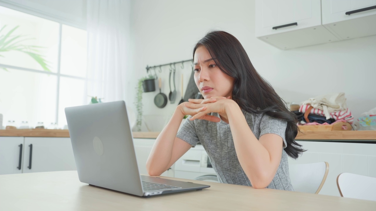 疲惫不堪的亚洲女性在家里用笔记本电脑工作。忙碌的员工女孩感到疲惫和头痛，因为工作很晚，工作文件在客厅的笔记本上。视频素材