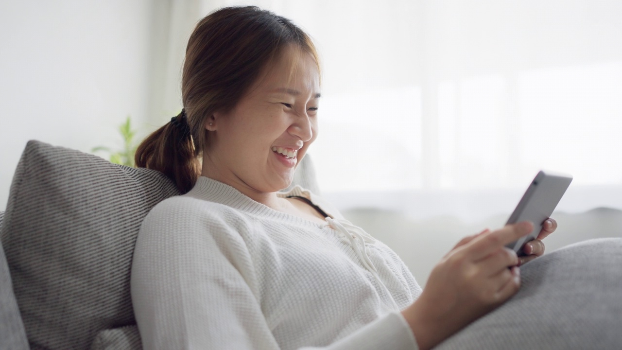 年轻女子在家里的沙发上玩电子平板电脑，玩得很开心。视频下载