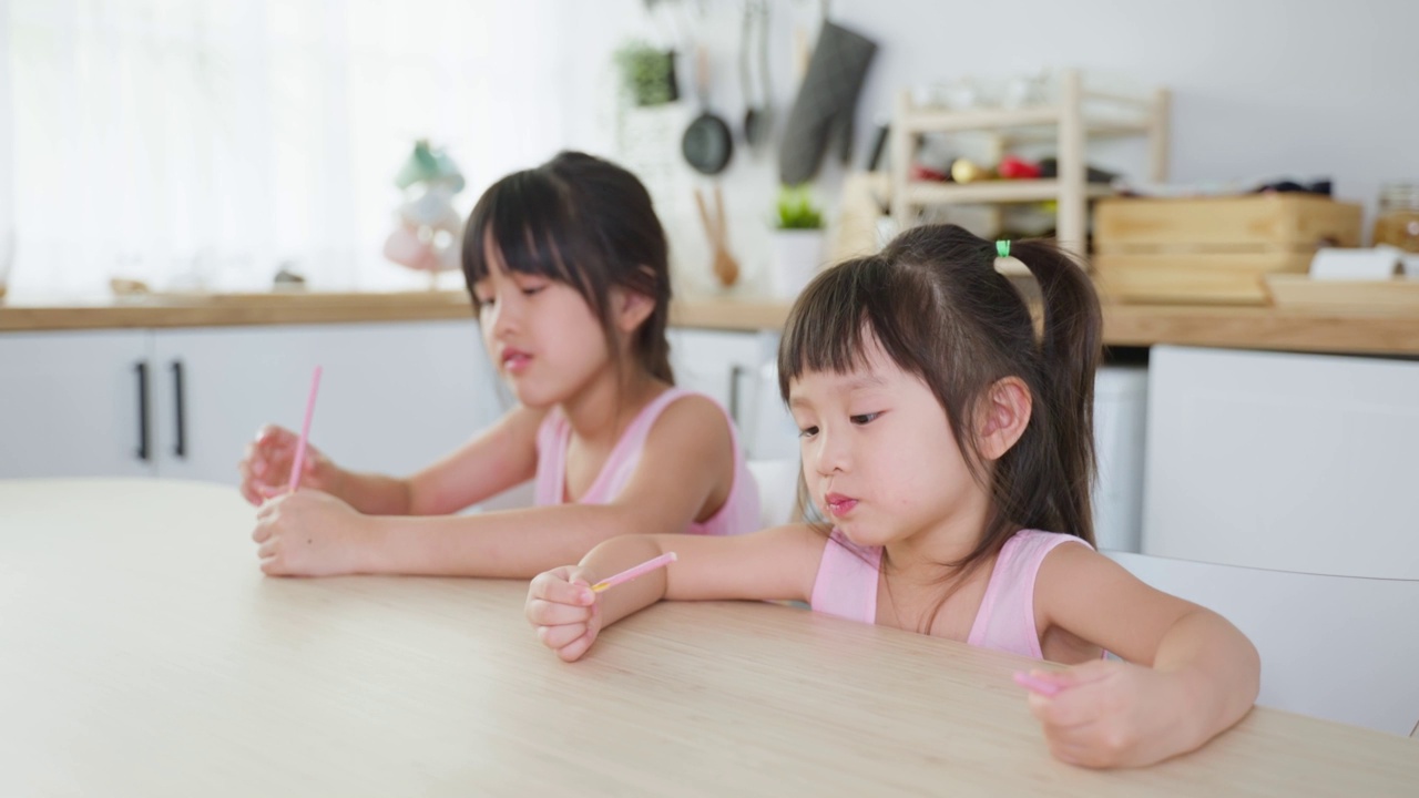 亚洲两姐妹甜甜的小女孩喜欢吃快餐、薯片。饥饿的学龄前女孩坐在厨房的桌子上，用手把零食放进嘴里。对儿童来说，美味而不健康的食物的概念。视频素材
