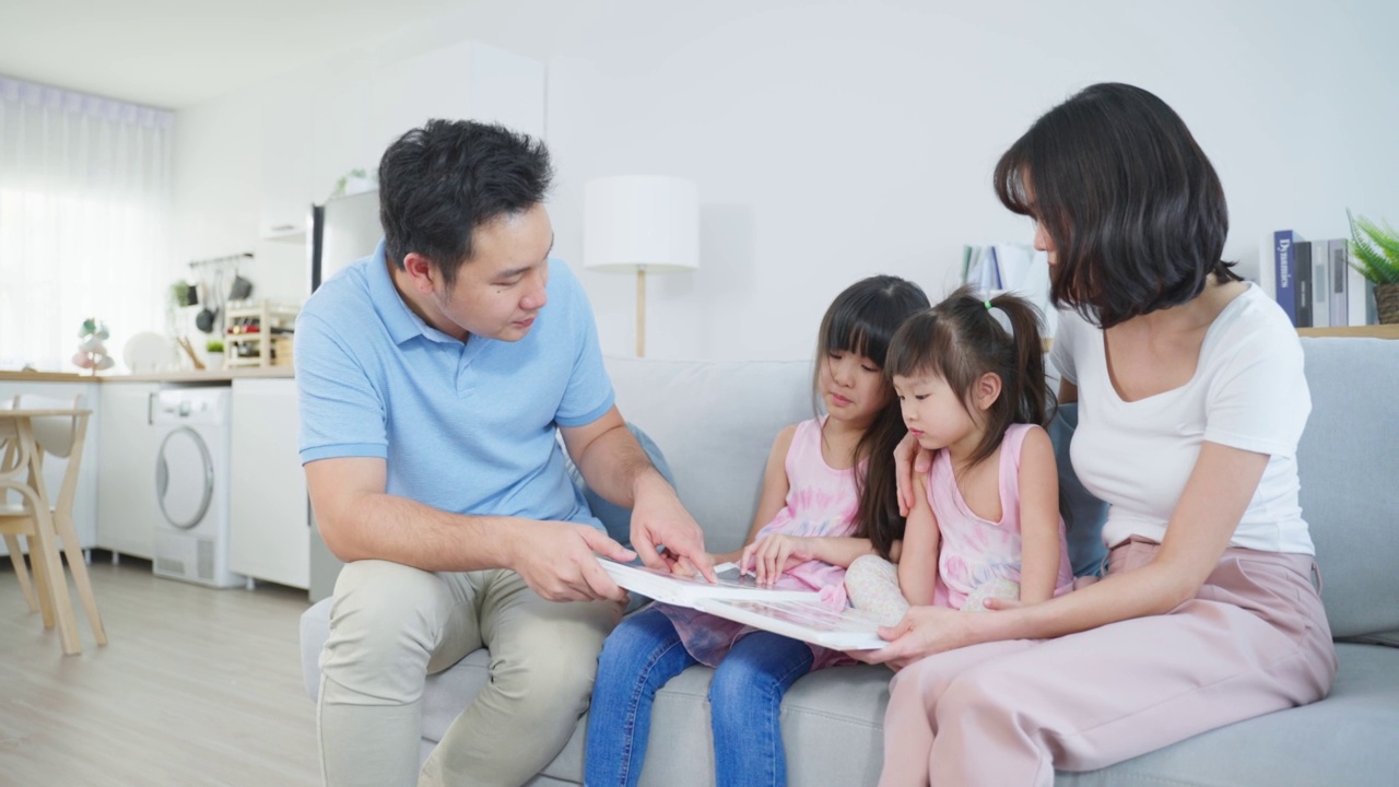 亚洲家庭看着旧相册上的照片回忆过去。年轻美丽的夫妇与年幼的女儿坐在一起翻阅相册，享受回忆关系的记忆。视频素材