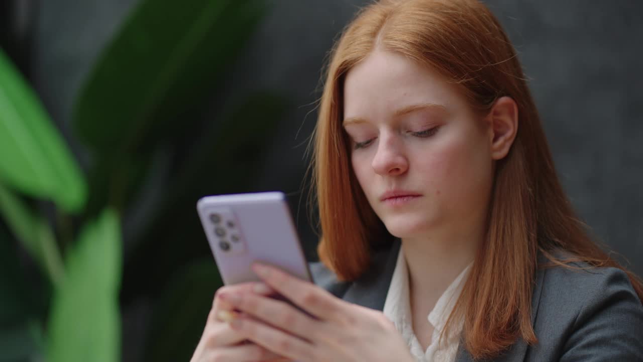 漂亮的年轻女商人在办公室使用电话。女人使用智能手机，靠在窗口发短信，发邮件，计划会议，在线网络，浏览手机信息视频素材