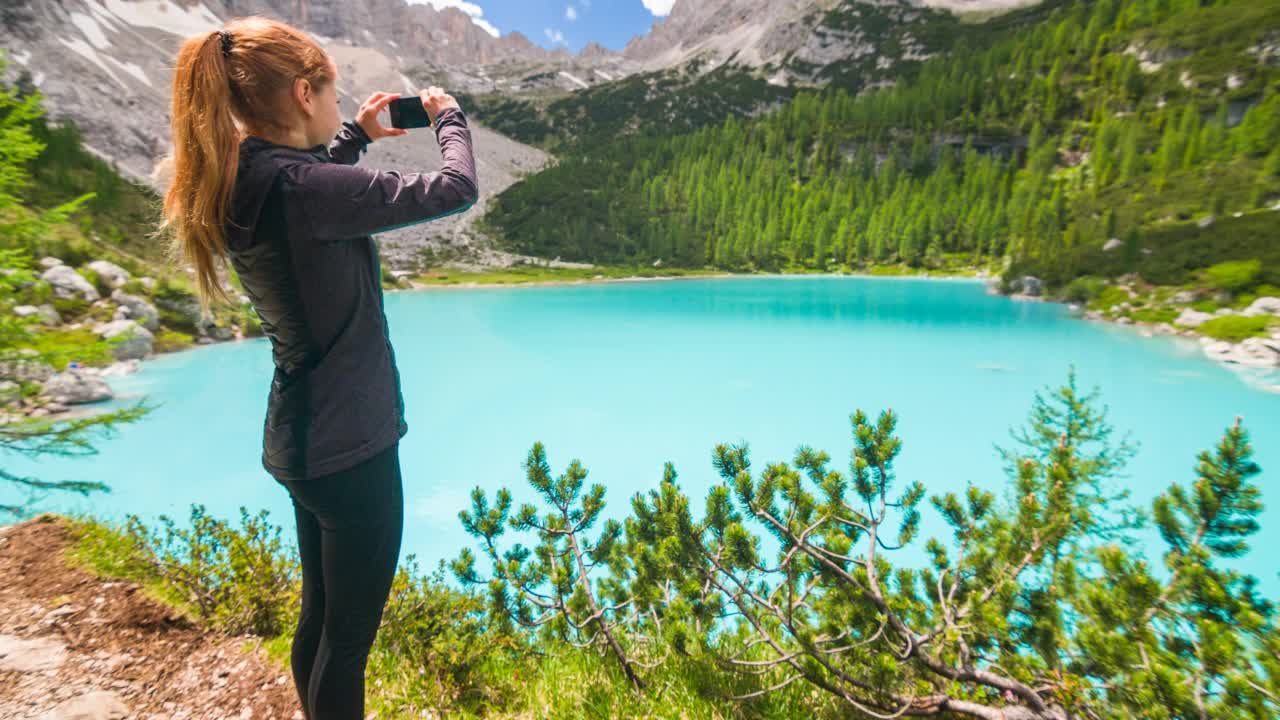 女游客在绿松石山湖欣赏惊人的景色，用智能手机拍照，山在背景视频素材