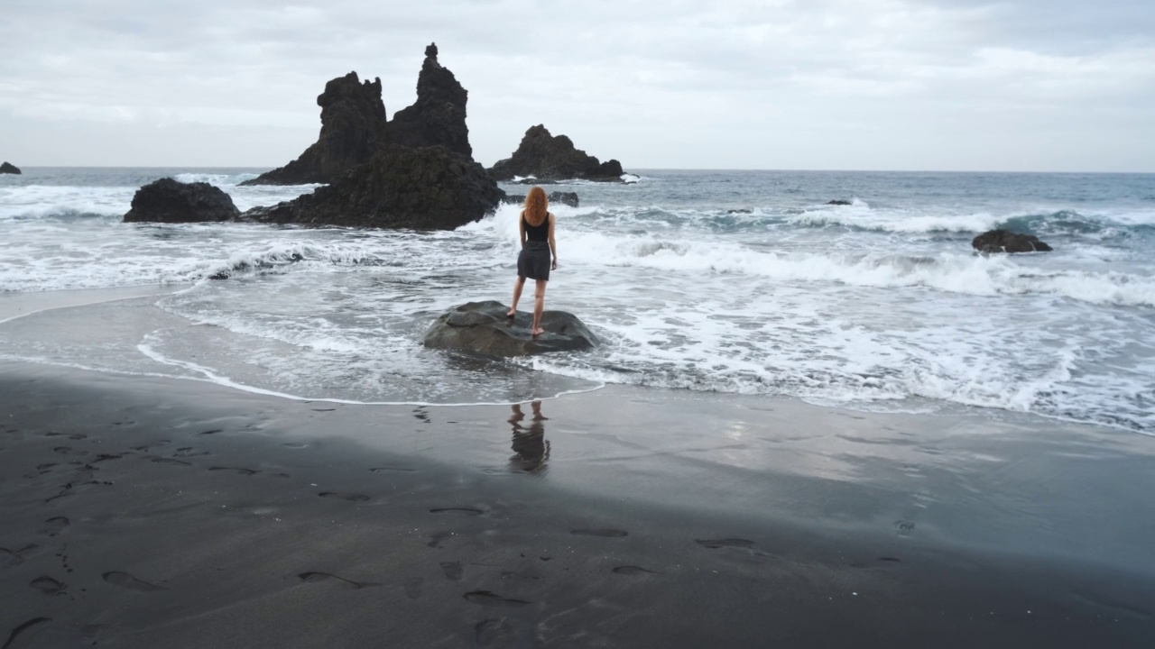 在加那利岛北部的特内里费岛，穿着裙子的女人站在黑火山黑沙滩贝尼霍岸边的一块大岩石上视频素材