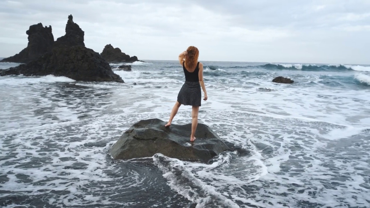 在加那利岛北部的特内里费岛，穿着裙子的女人站在黑火山黑沙滩贝尼霍岸边的一块大岩石上视频素材