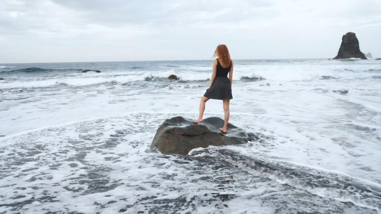 在加那利岛北部的特内里费岛，穿着裙子的女人站在黑火山黑沙滩贝尼霍岸边的一块大岩石上视频素材