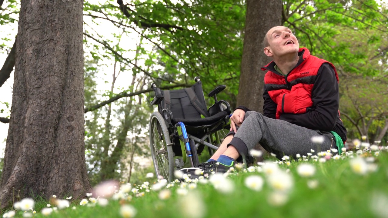 一个瘫痪的年轻人坐在轮椅旁边的草地上，戴着一朵雏菊，微笑着享受大自然的肖像视频素材