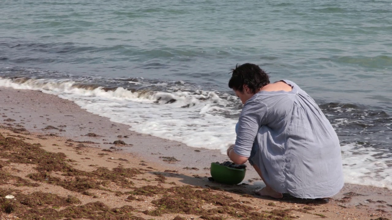 海滩上的女人在海浪中用海水洗碗中的贻贝。海景度假视频素材