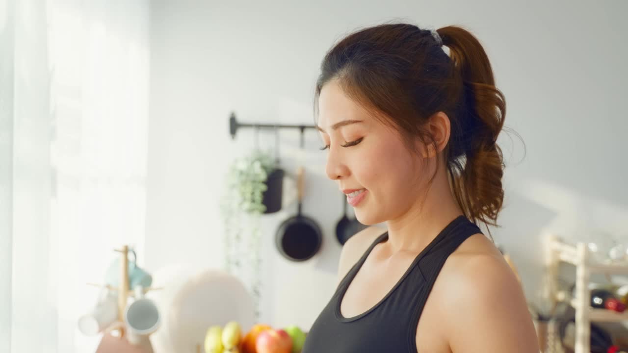 穿着运动服的亚洲美女运动后在家喝水。年轻的口渴活跃的运动女孩喝干净的天然矿物在杯子锻炼后，在家里的厨房保健。视频素材