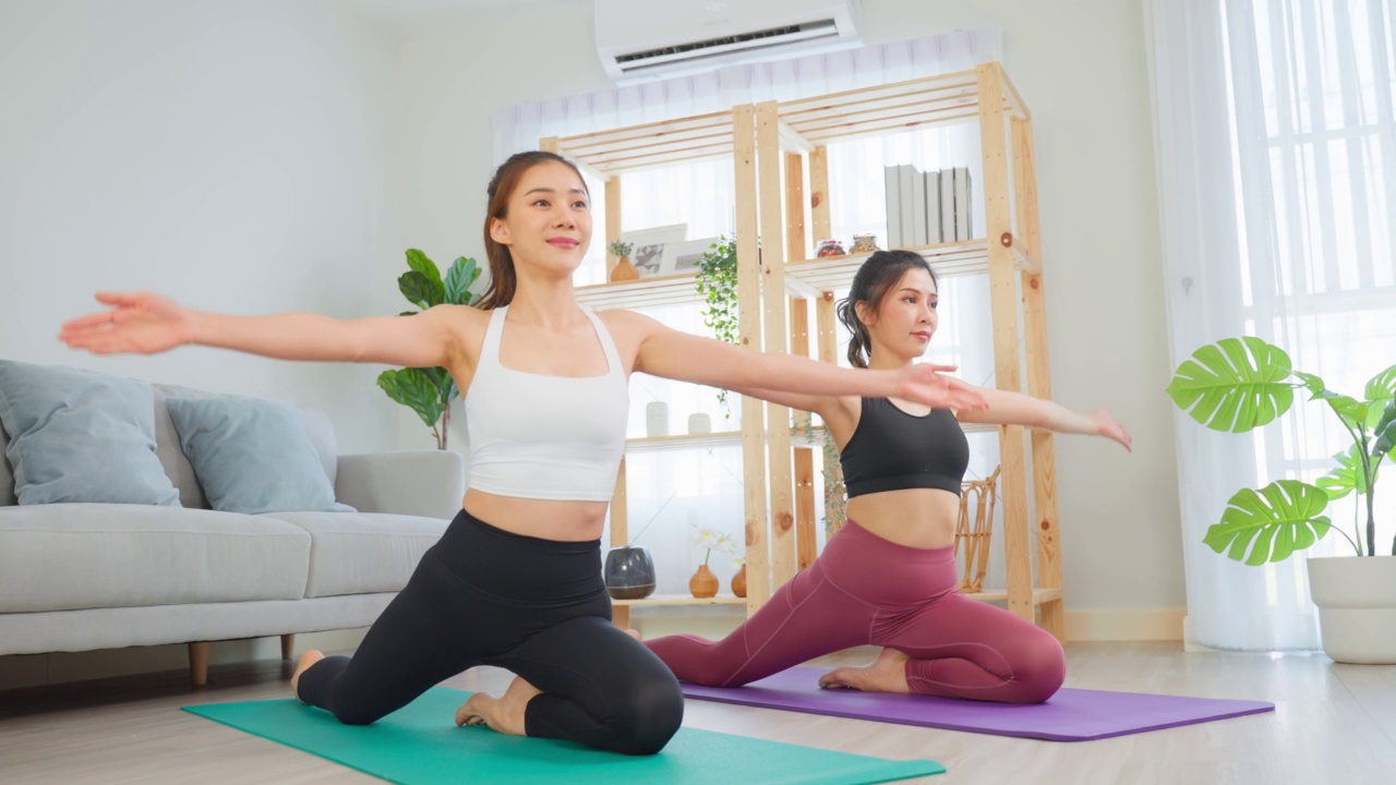 亚洲活跃的女性朋友在家里一起做瑜伽普拉提锻炼。漂亮的女孩穿着运动服，在家里的客厅里用垫子花空闲时间健身保健。视频素材