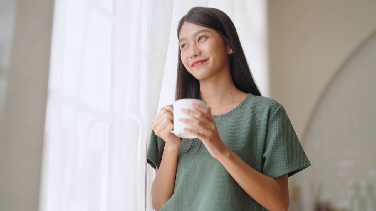 年轻的亚洲妇女站在窗边，拿着杯子在家里的卧室，她早上醒来后喝牛奶视频素材