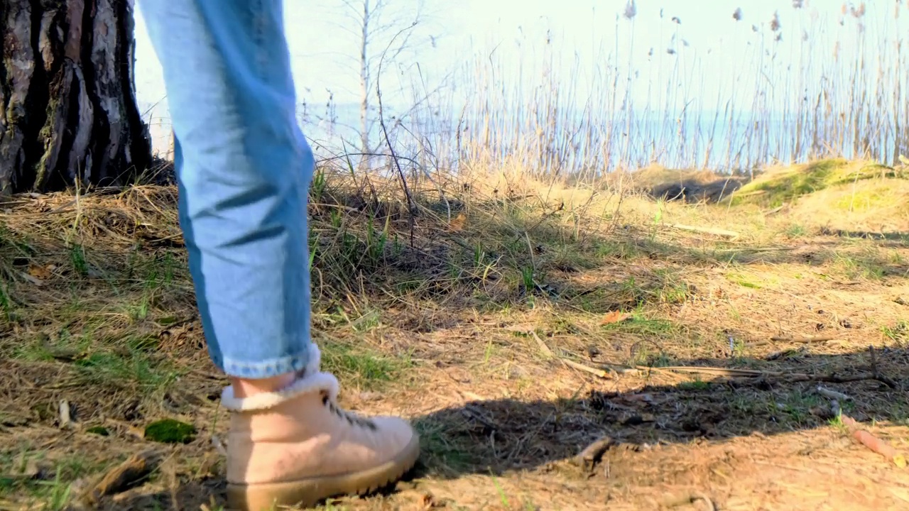 徒步旅行。镜头跟随在春天的森林里，穿着靴子的女人的脚，地面上的球果和掉落的针。一个人走近以河流或湖泊为背景的悬崖。慢镜头,4 k视频素材