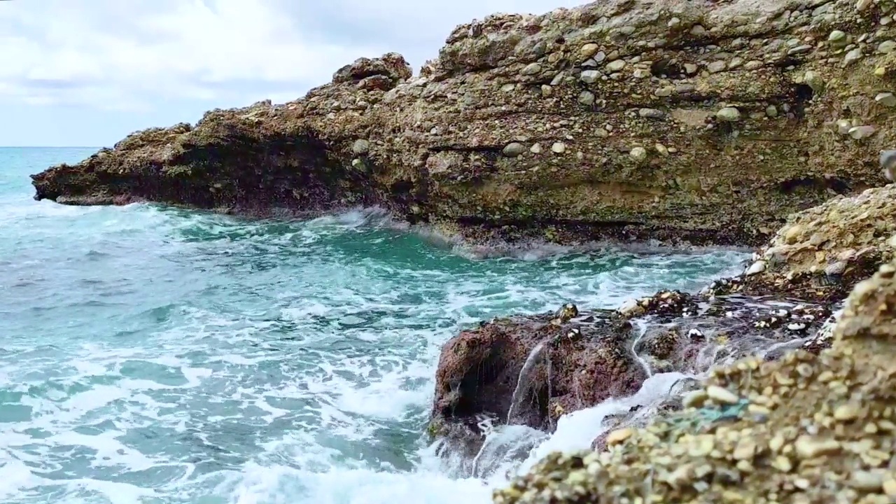 海浪拍打着海岸上的石头。蓝色的大海，淡淡的暴风雨，白色的浪花，晴朗的天气。视频下载