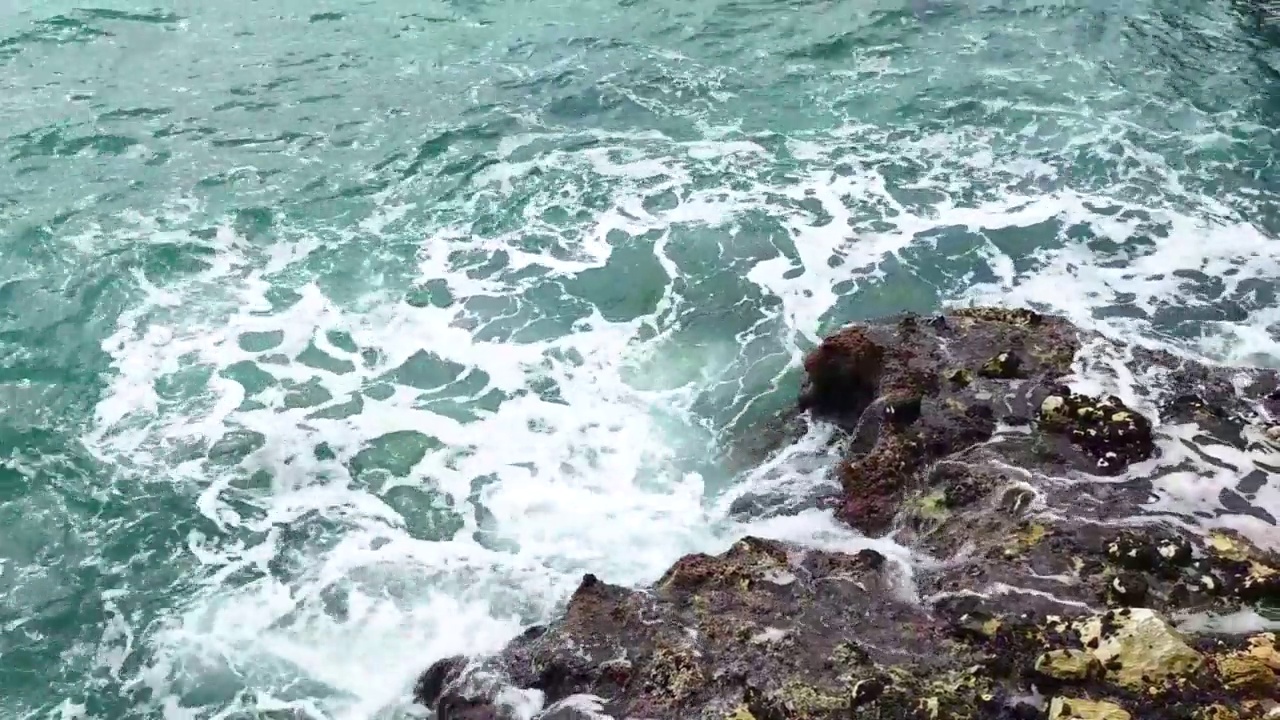 海浪拍打着海岸上的石头。蓝色的大海，淡淡的暴风雨，白色的浪花，晴朗的天气。视频下载