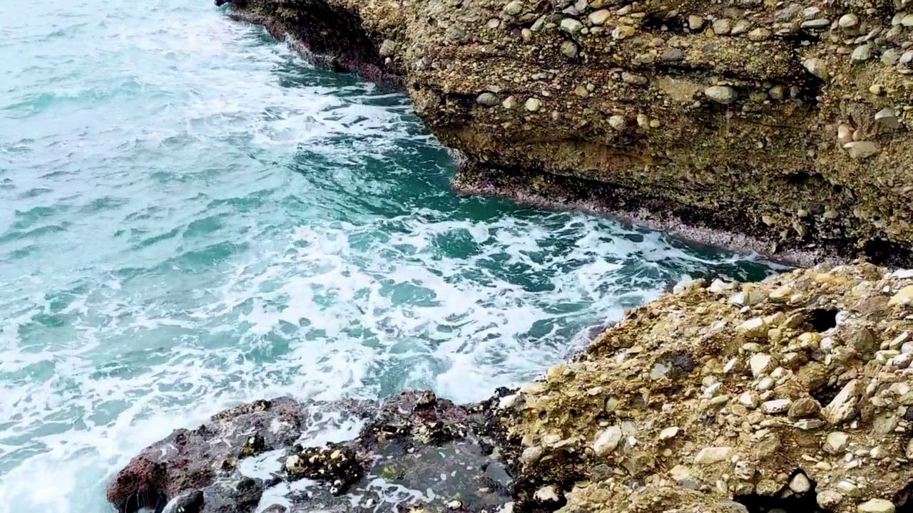 海浪拍打着海岸上的石头。蓝色的大海，淡淡的暴风雨，白色的浪花，晴朗的天气。视频下载