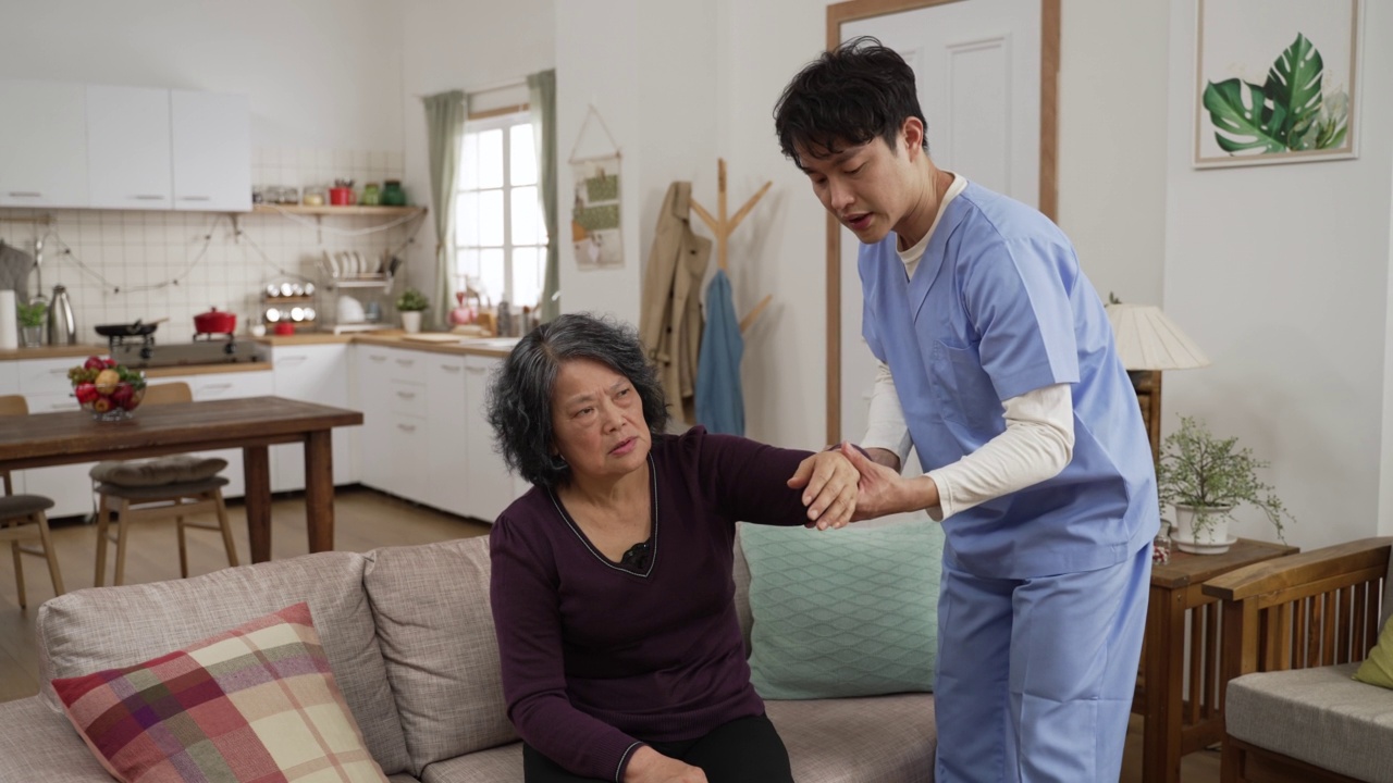 亚洲男性理疗护士在家里的客厅帮助老年女性做康复运动。当她因不适而哭泣时，他慢慢地抬起她无力的手臂视频素材