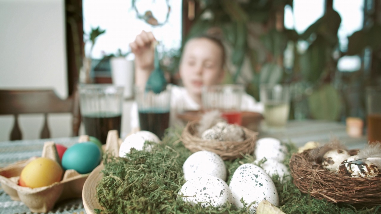 一个五岁的金发女孩正在享受给复活节彩蛋着色的乐趣。女孩把鸡蛋浸入一杯颜料中。温馨的家庭氛围和复活节装饰。股票视频。视频下载