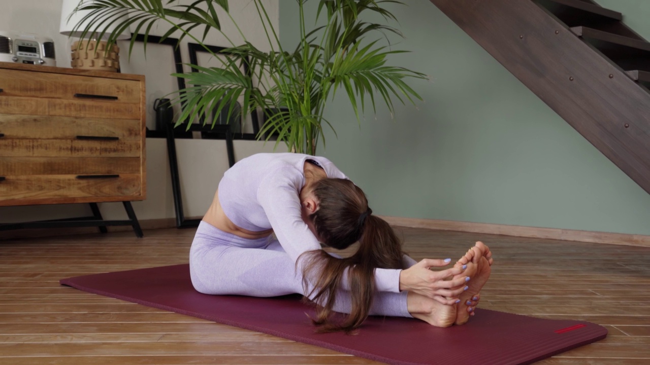 年轻女子在家伸展身体。一位身穿紫色西装的女士坐在瑜伽垫上，弯腰把脚向自己拉，伸展腿部肌肉视频素材