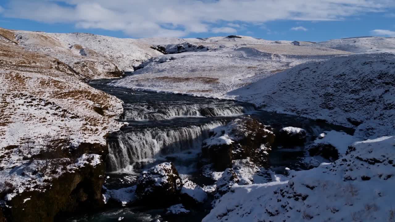 在阳光明媚的冬天，冰岛南部海岸Skógá村附近，Skógá河附近Fimmvörðuháls小道的水瀑布，岩石峡谷周围被白雪覆盖的草地。视频素材