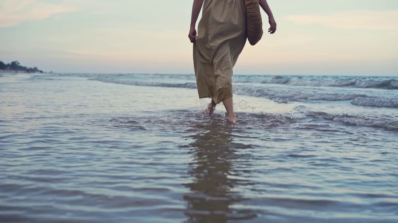 女人的脚沿着海浪走在沙滩上。漂亮女人在海边冲浪。夏日假期里，浪花和泡沫的慢动作。日落时穿着漂亮衣服的女人视频素材