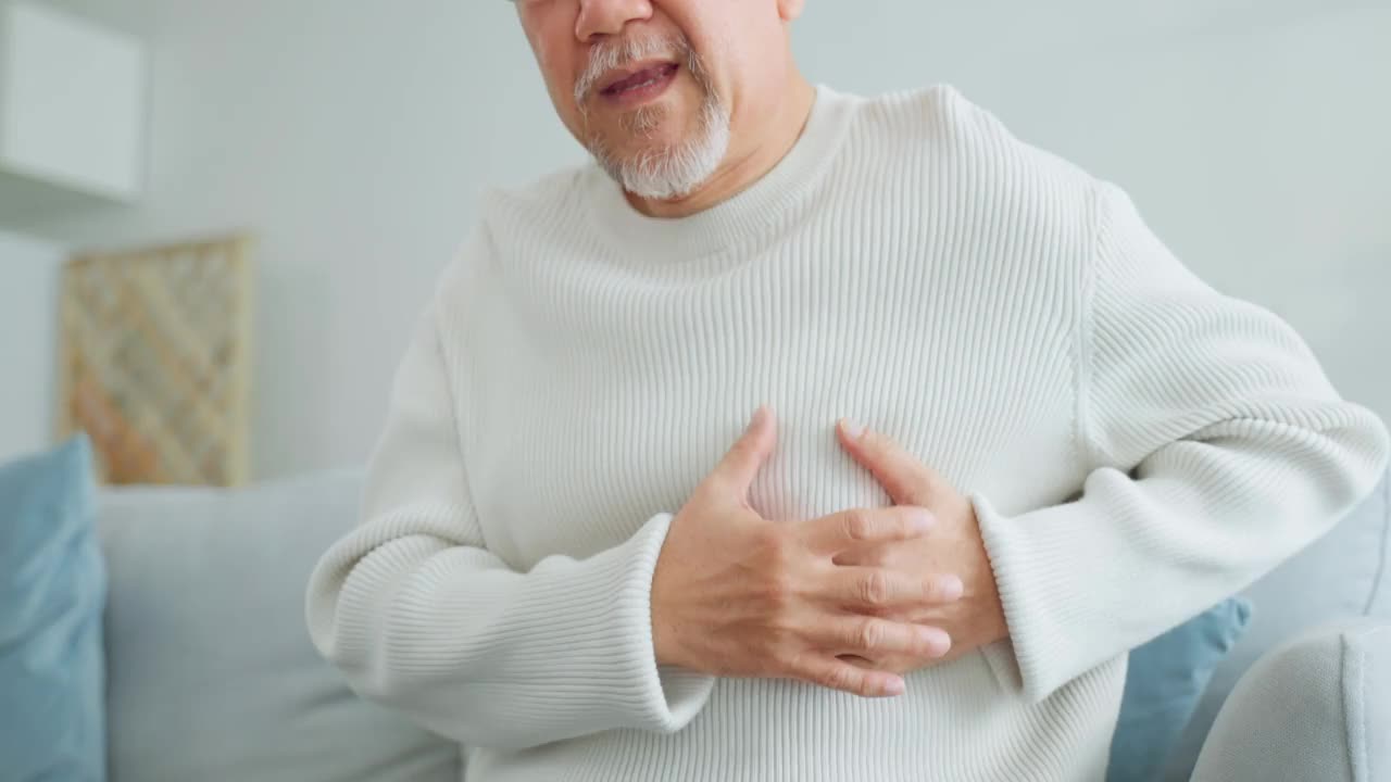 亚洲老年男子胸部疼痛，感觉心脏病发作。有魅力的老年病人坐在客厅的沙发上，因剧烈疼痛而捂着胸口呼吸困难。视频素材