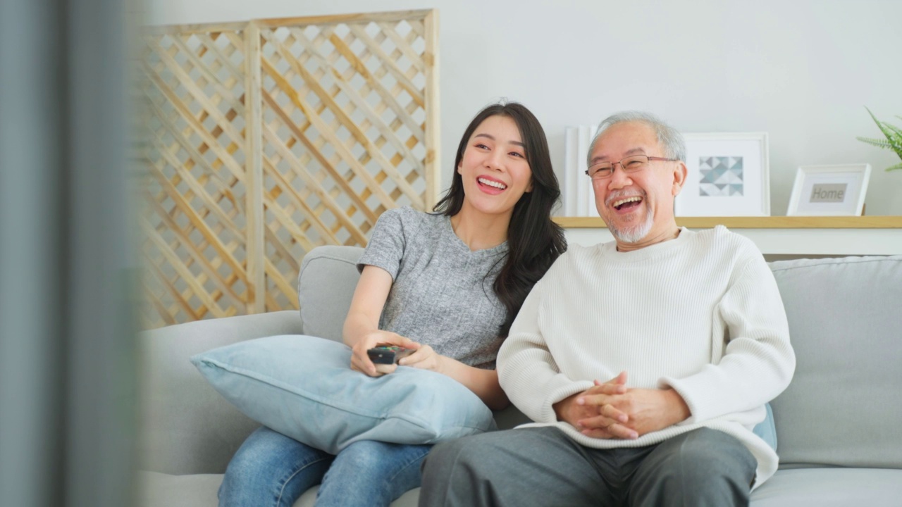 亚洲年长的父亲和女儿一起在家里看滑稽电影。慈祥的老爷爷和美丽的女孩孙女坐在沙发上，欣赏电视上的喜剧节目，在客厅里度过时光。视频素材