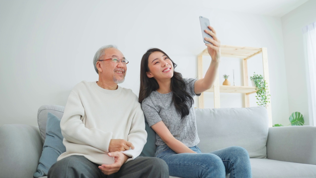 亚洲可爱的家庭，女儿用电话与资深父亲视频通话。年轻漂亮的女孩和上了年纪的老男人坐在沙发上享受网上交流，在家里的客厅里一起聊天视频素材