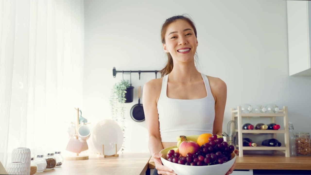 亚洲有吸引力的活跃的女人拿着一碗水果在家里的厨房。活跃的运动女孩的肖像在运动服享受吃苹果健康食品为健康锻炼后，看在房子里的相机。视频素材