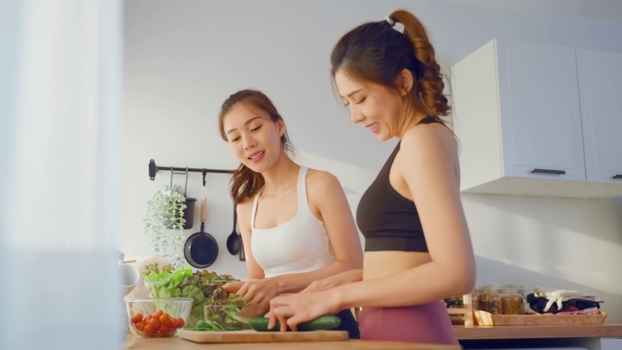 两个穿着运动服的亚洲年轻女子在厨房里做沙拉。活泼漂亮的女孩姐姐感到快乐，喜欢吃蔬菜健康的食物来减肥，在家里的保健视频素材