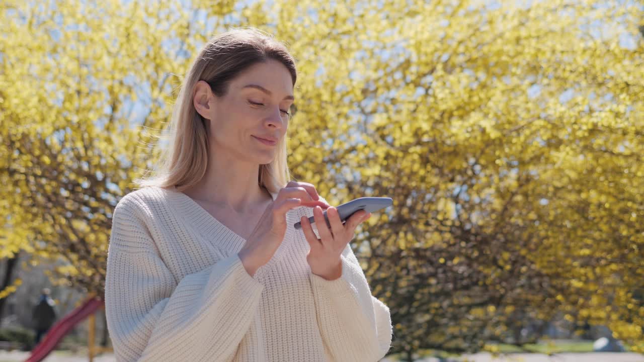 穿着白色毛衣的漂亮女人在一个温暖的阳光明媚的日子里，在公园的一棵黄叶树附近用手机录音发送语音语音信息。视频素材