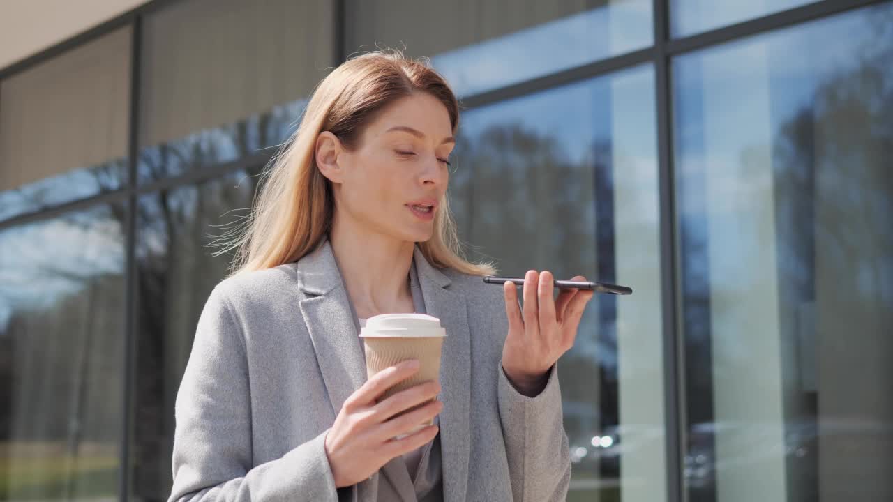 女商人在智能手机上发送语音信息，拿着早上的咖啡，站在现代办公大楼附近，与移动助理交谈。正装女商人使用手机应用程序。视频素材