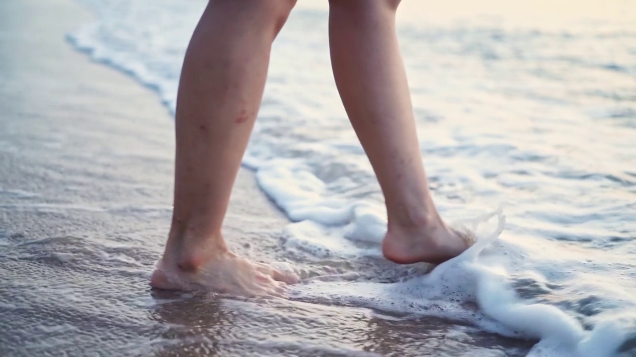 女性的腿和脚沿着海浪走在沙滩上。受伤的腿女人走在海边冲浪。水花和泡沫的慢动作。女孩在日落或日出时在海洋里。视频素材