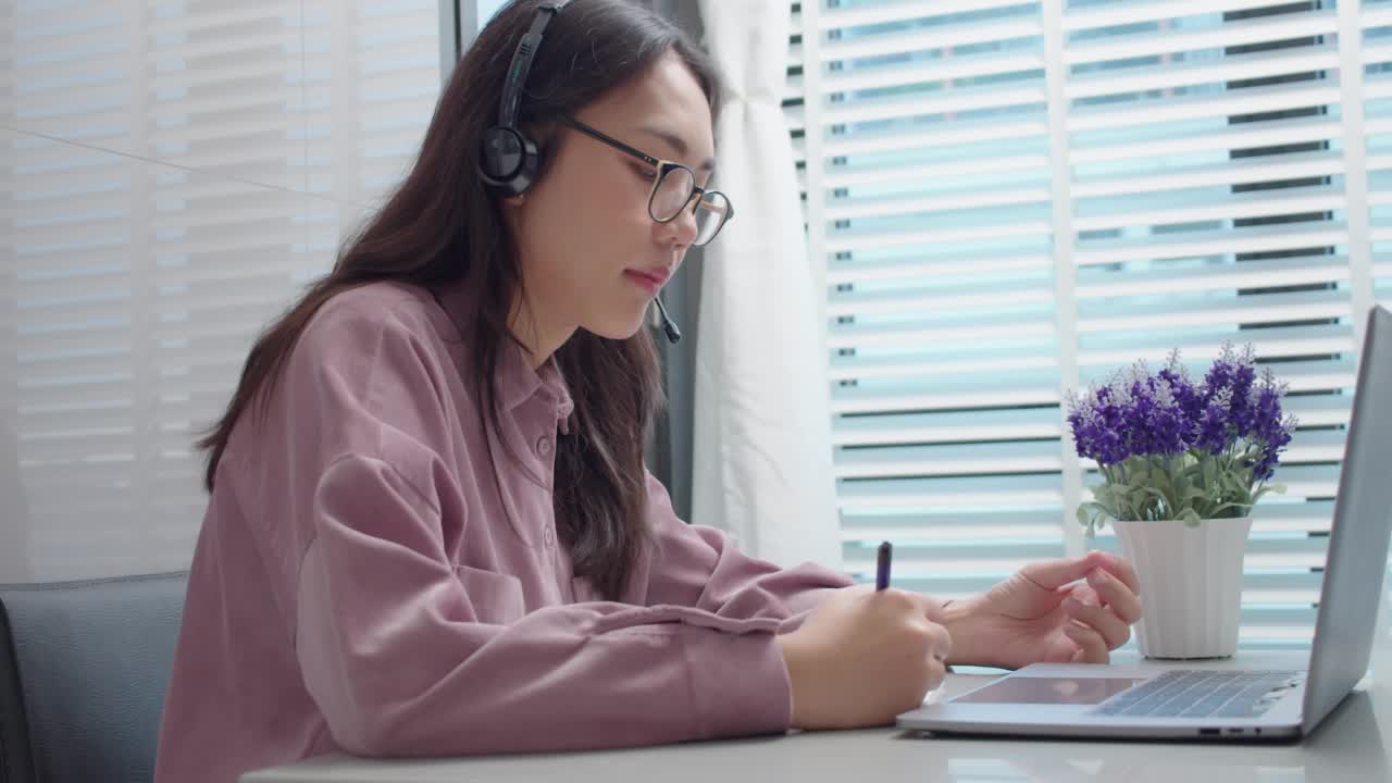 亚洲女学生在家桌上用笔记本电脑在线学习。女性戴着耳机，看着笔记本电脑说话，解释和写笔记。在家学习。在线学习的概念。视频素材