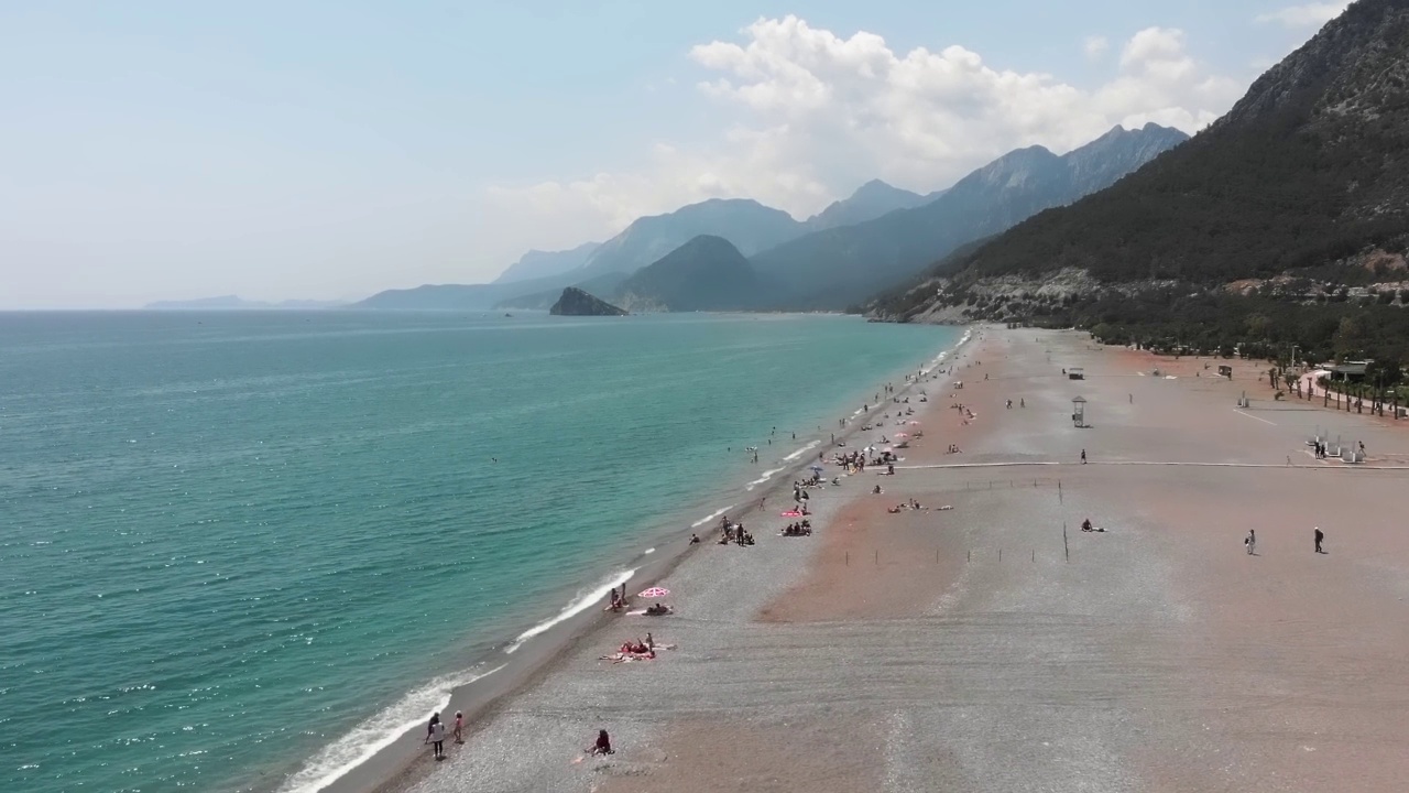 宽阔的沙滩与山脉背景。地中海海滨。夏天人们在海里游泳，在沙滩上晒日光浴。夏季海滩度假概念。旅游目的地视频素材