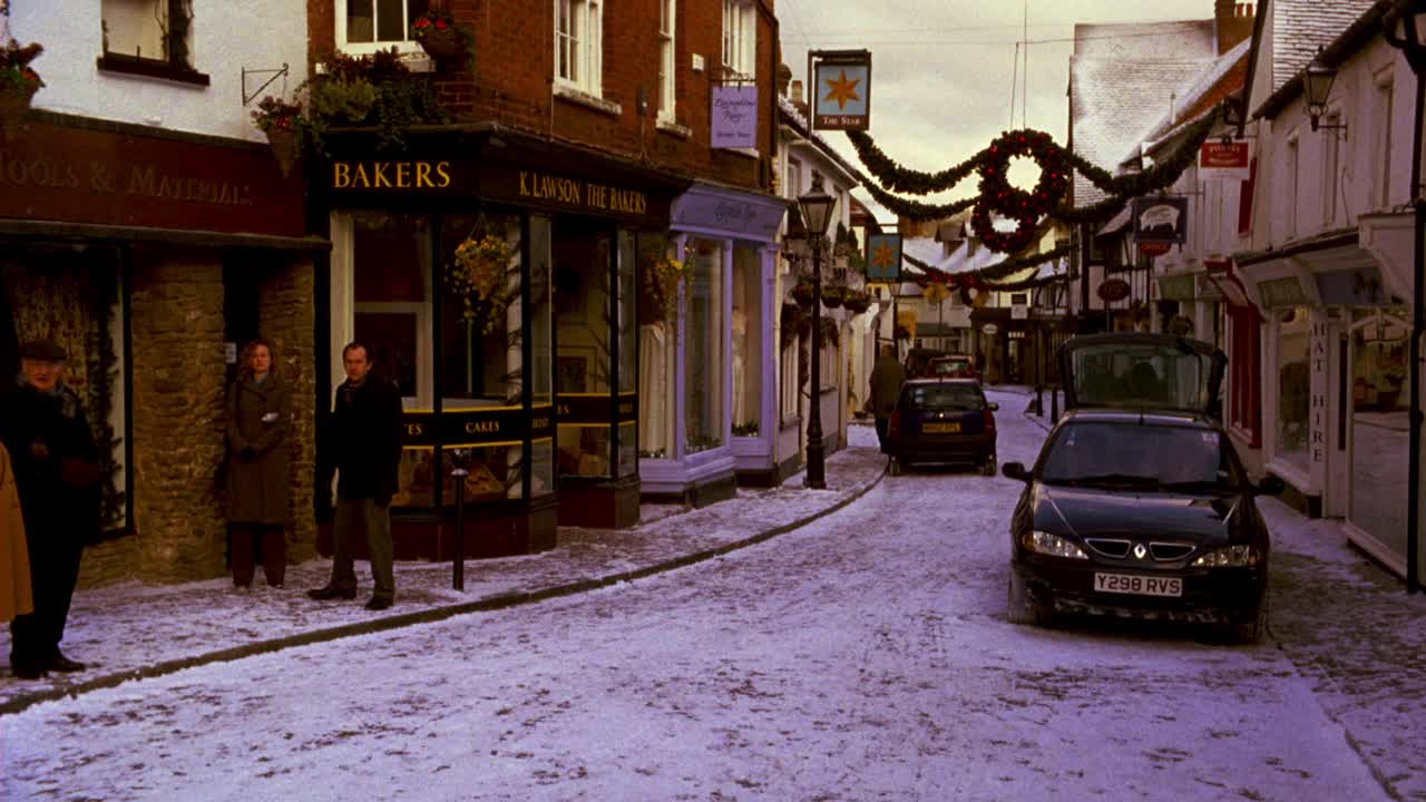 宽角度的雪覆盖街道通过英语村的店面，包括面包店。村民，老夫妇过马路，男人骑自行车。街上挂满了圣诞节用的装饰花圈。视频素材