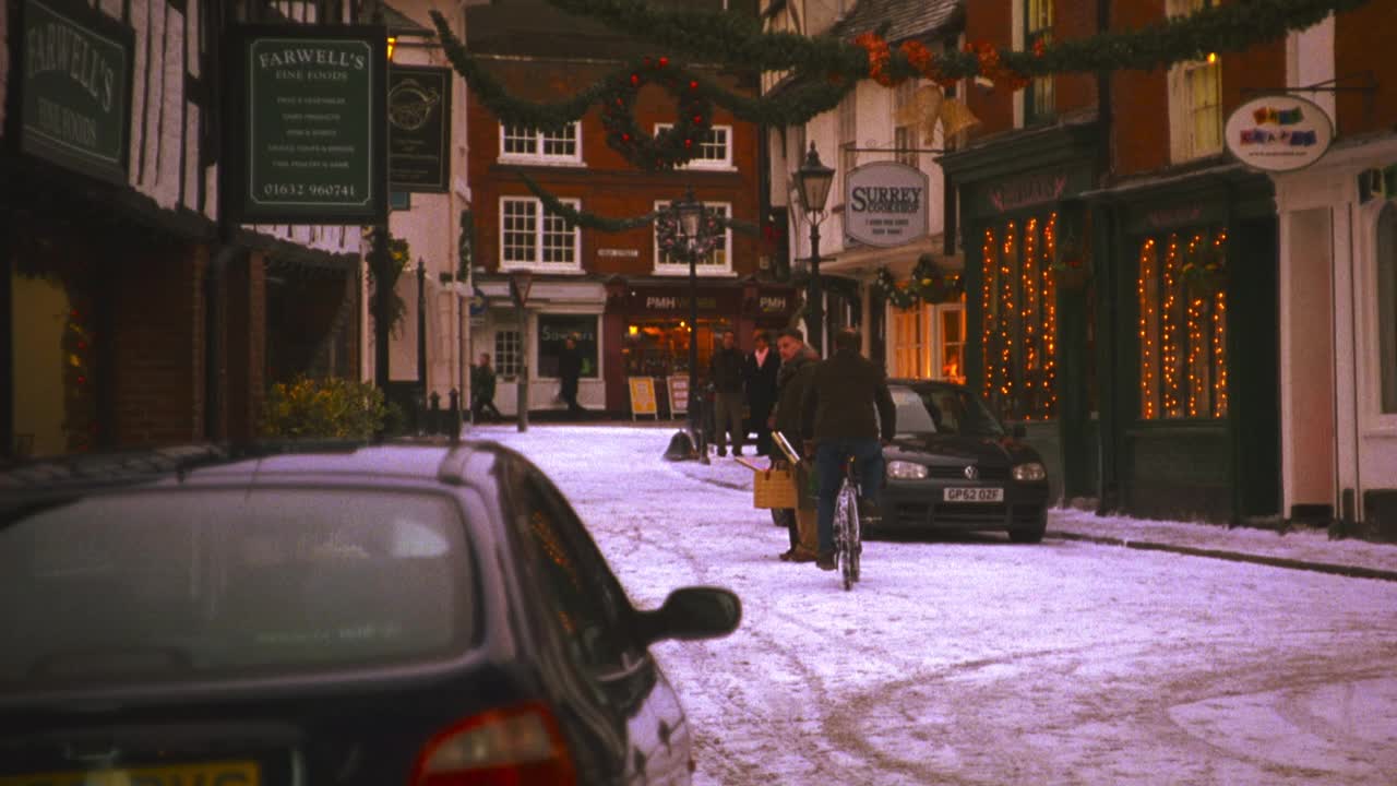 广角的英国村庄，狭窄的白雪覆盖的街道和店面，包括“告别的美食”。圣诞彩灯、装饰品和花环悬挂在建筑物上。村民们，男人和女人走在人行道上，男人骑着自行车视频素材