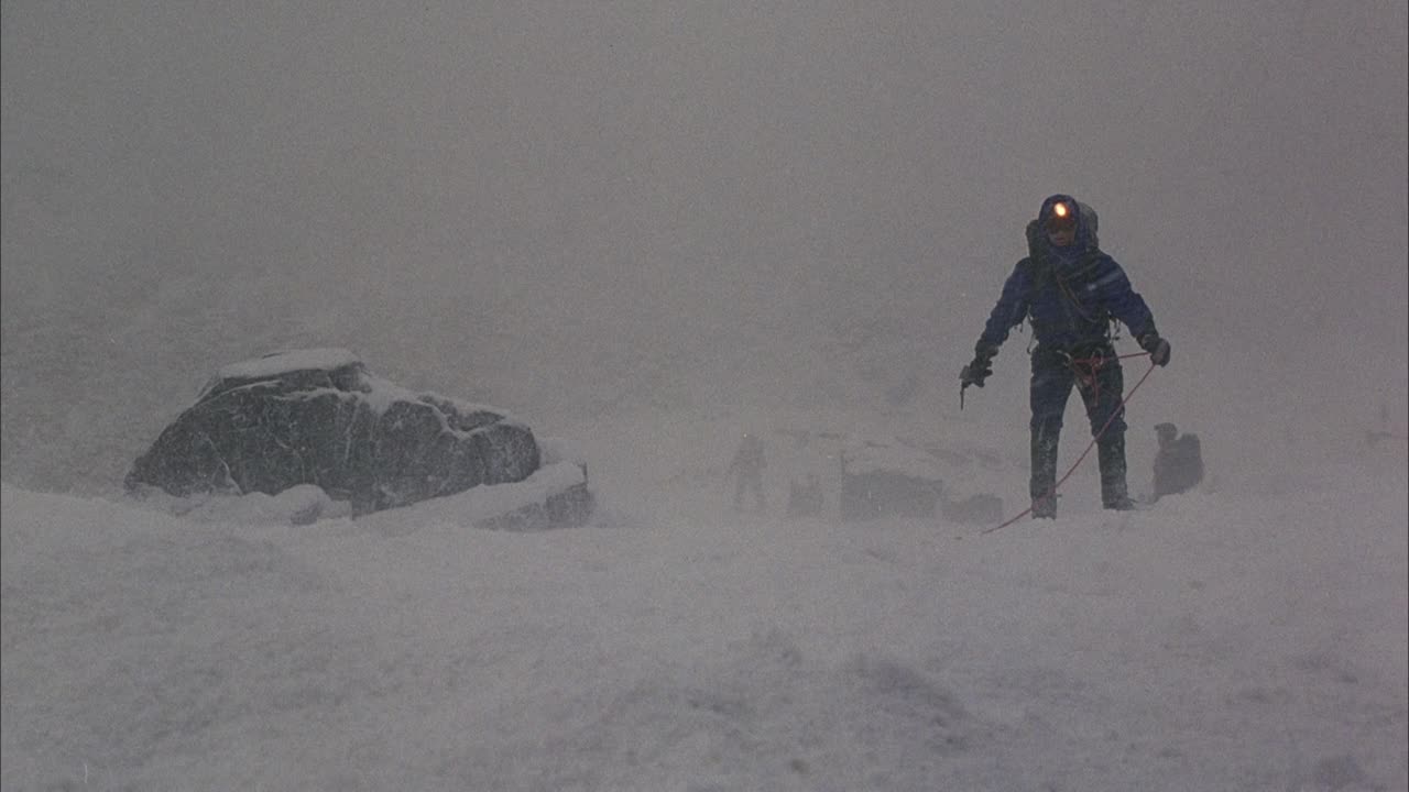 中等角度的登山者在暴风雪中站在雪中。看到第一个攀登者拿着向左延伸的红绳子，攀登者挣扎着走向坡。绳索拉紧并缠绕在第一个攀登者的腿上。看攀岩者向后拖视频素材