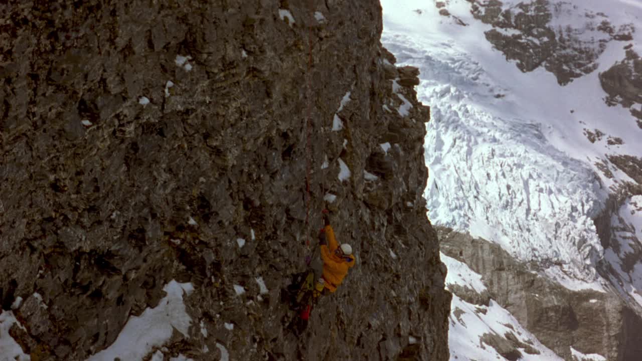 中等角度的男性登山者，抓住绳子，爬上布满雪的岩石山坡或悬崖。看到攀登者左侧的背包随着攀登者向上移动而向上抖动。在背景中看到被雪覆盖的地面和巨石。视频素材