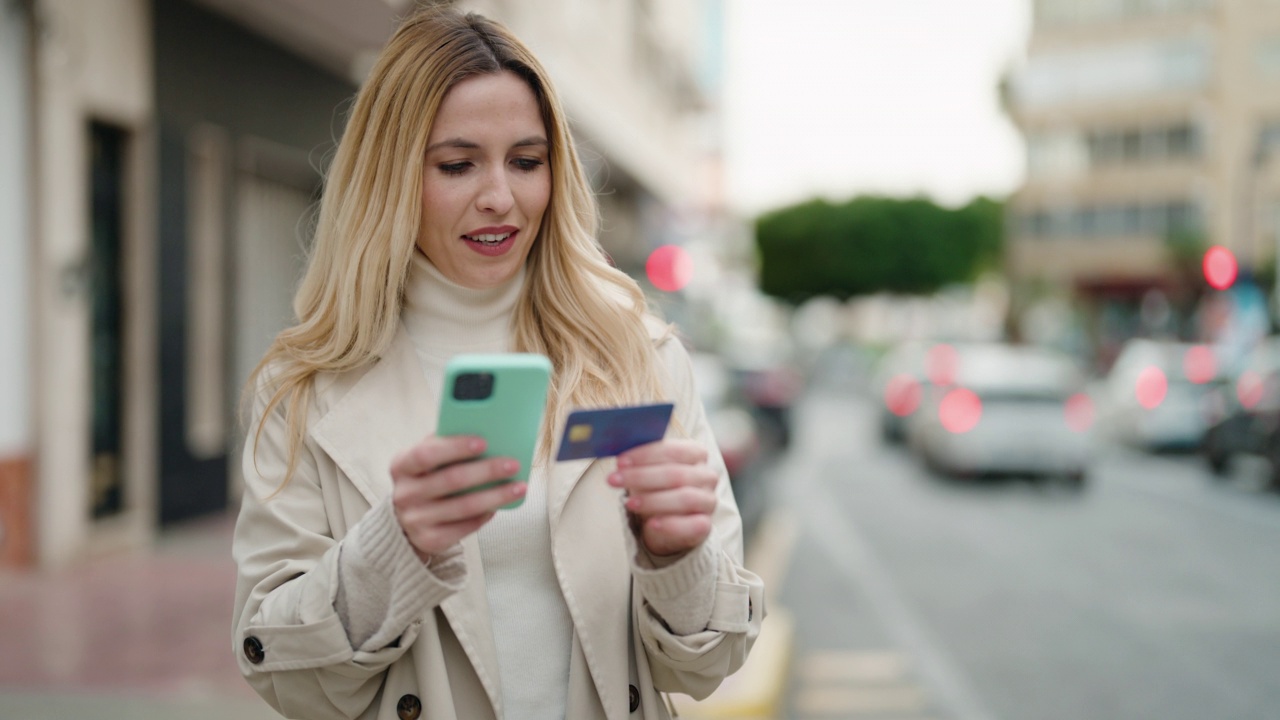 年轻的金发女郎在街上用智能手机和信用卡视频素材