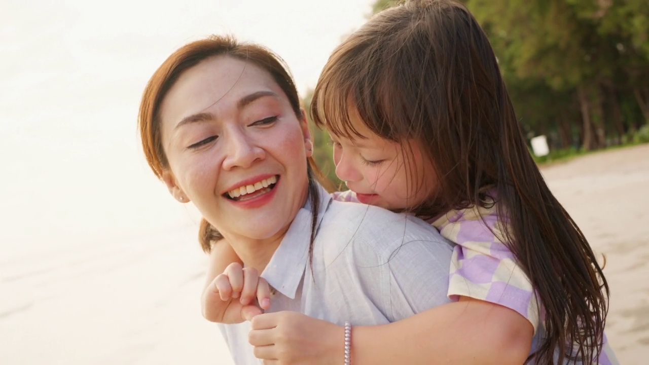 4K亚洲母亲背着小女儿，在夏天的夕阳下走在热带海滩上。视频素材