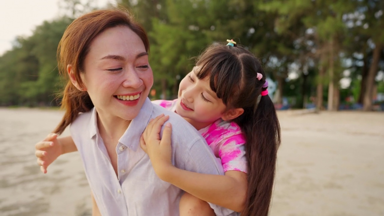 4K亚洲母亲背着小女儿，在夏天的夕阳下走在热带海滩上。视频素材