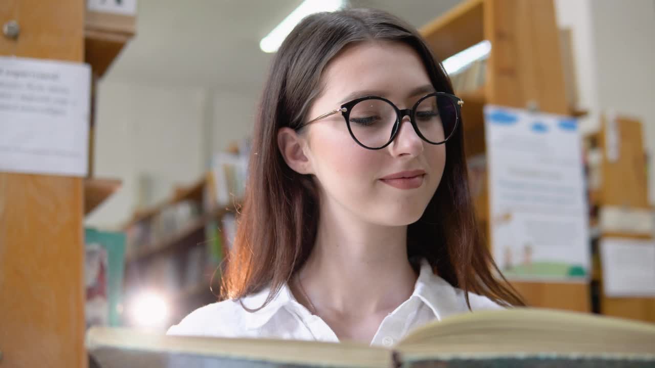 一个戴眼镜微笑着的年轻女孩在大学图书馆看书视频素材