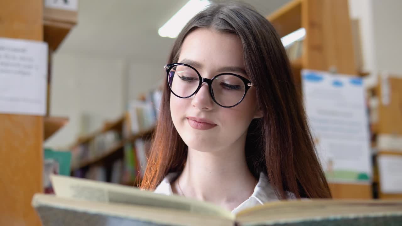 一个戴眼镜微笑着的年轻女孩在大学图书馆看书。一个有思想的年轻学生站在图书馆里看书视频素材