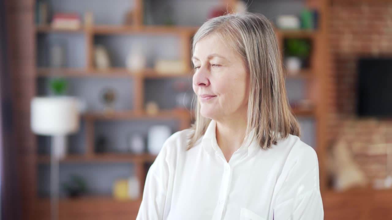 肖像一个年长的灰色头发的女人看着相机和微笑。室内。在现代家庭或办公室。特写美丽幸福的老生意女交叉双臂的脸视频素材