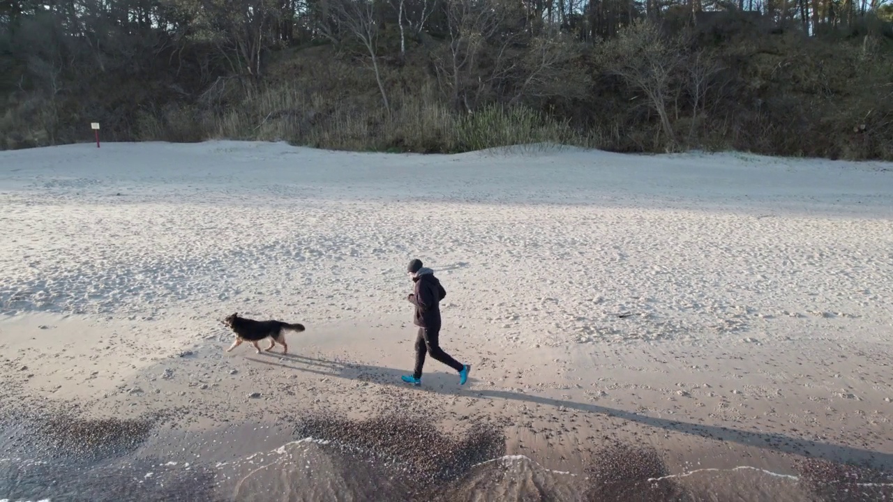 日出时，穿着衣服的人带着他忠实的狗在空旷的海滩上奔跑。人与动物之间的友谊。视频素材