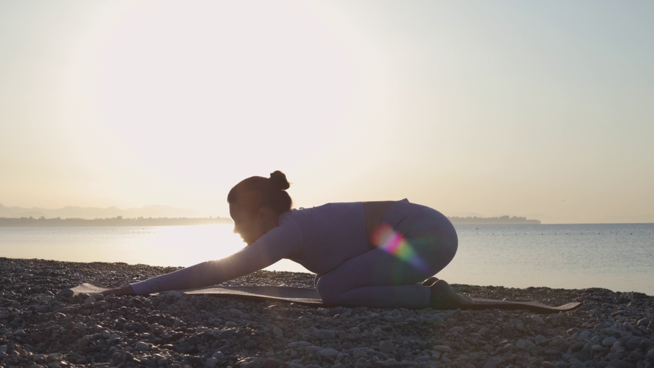 在日出或日落时，健康的年轻女子在平静的海面附近的海滩上练习瑜伽视频素材