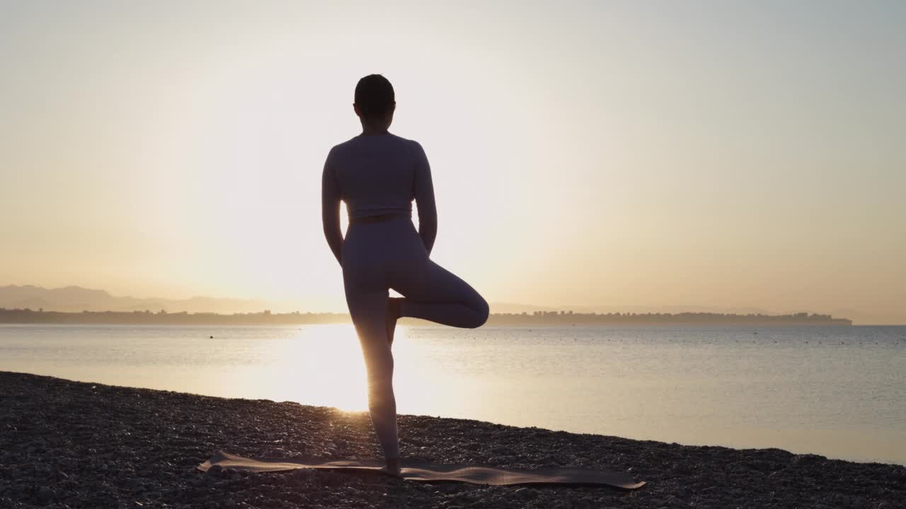 在日出或日落时，健康的年轻女子在平静的海面附近的海滩上练习瑜伽视频素材