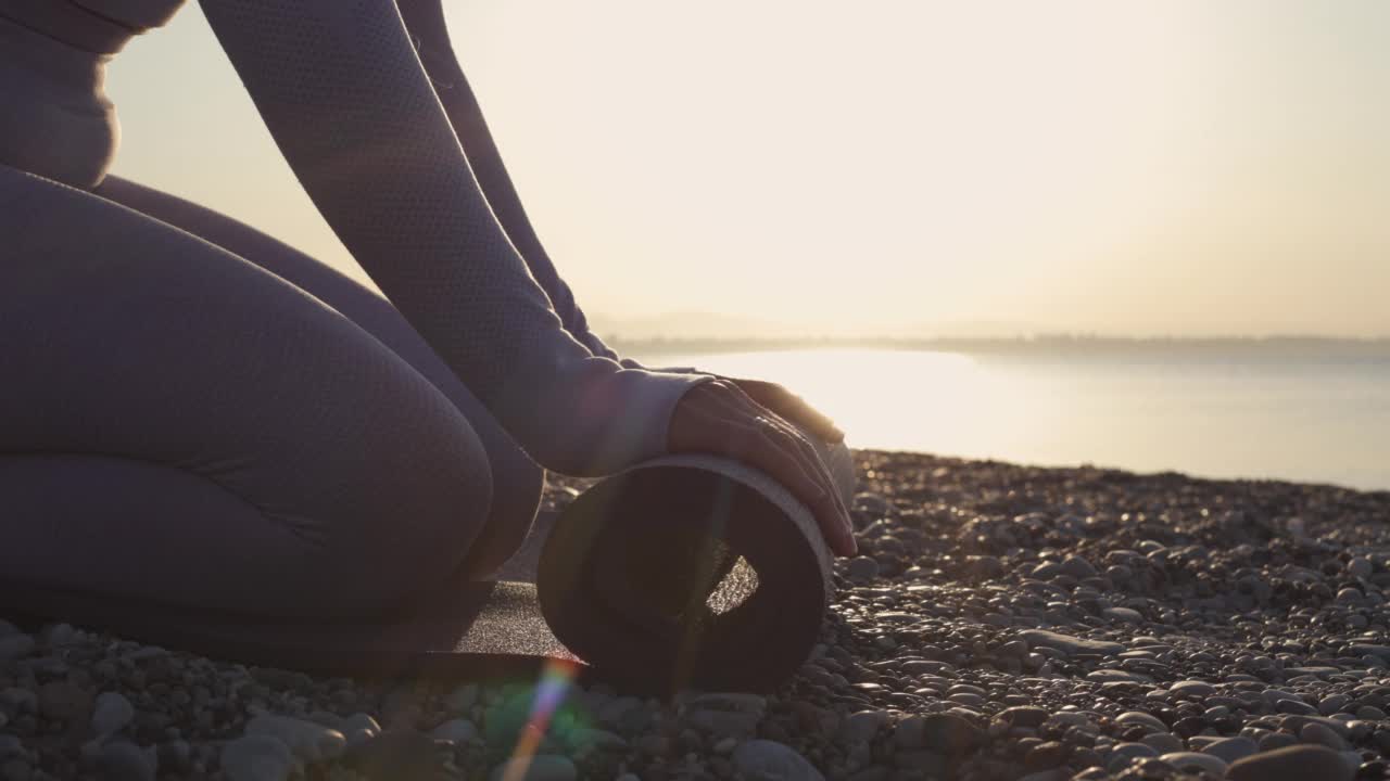 在日出或日落时，健康的年轻女子在平静的大海附近的海滩上进行瑜伽练习前滚动垫子视频素材