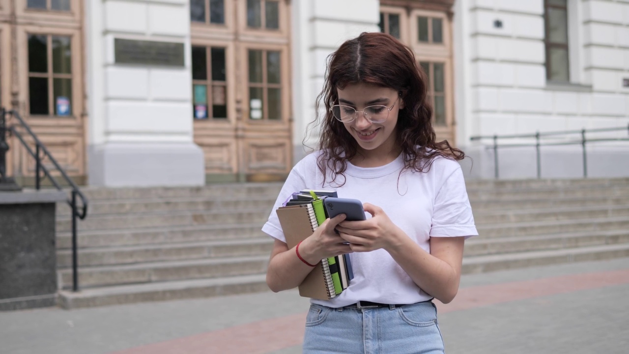 女卷发学生在白色t恤与书籍走在大学附近使用手机在校园户外。漂亮的女孩冲浪社交媒体，冲浪互联网，轻敲屏幕智能手机。视频素材