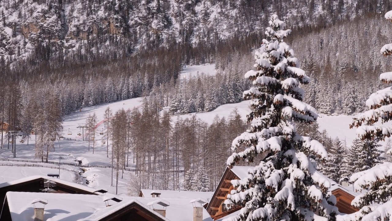 冷杉树和积雪的房子的屋顶在前景和滑雪斜坡和滑雪升降机在意大利阿尔卑斯山脉在大雪后充满雪视频素材